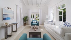 Un salón espacioso con un sofá blanco, un sillón azul y una mesa de centro. Hay un cuadro en la pared y una planta en maceta en la esquina.