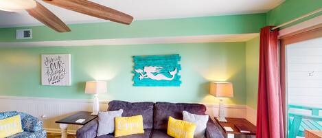 Een gezellige woonkamer met een grijze bank, twee gele kussens, een blauwgroene wanddecoratie en een plafondventilator.