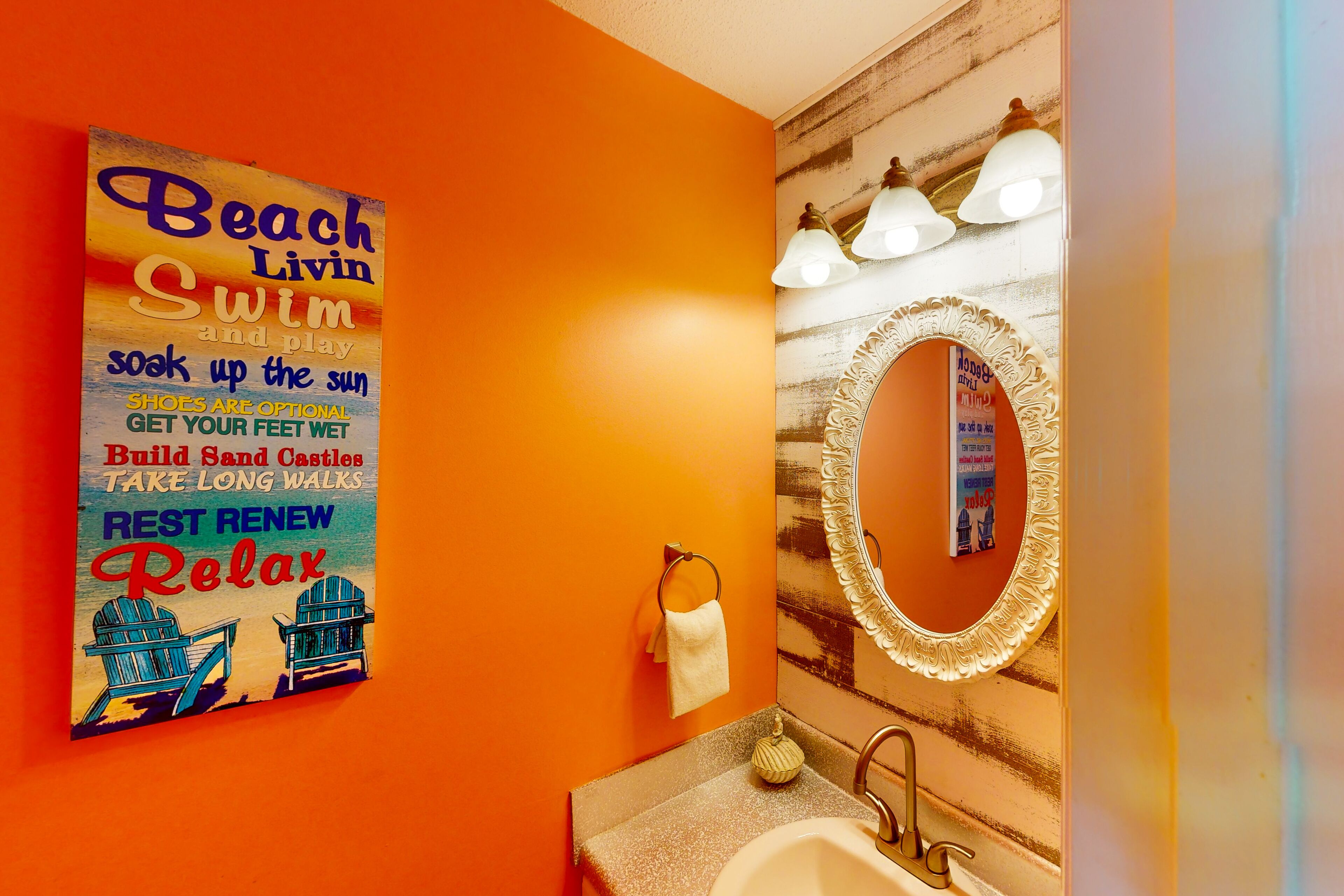 Un baño con una pared naranja, un espejo grande con marco decorativo y un lavamanos.
