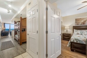 A bedroom with a bunk bed, a bed with floral bedding, a bedside table with a lamp, and a view of the ocean through large windows. - FREE Beach Service & Perks! Emerald Beach 732 Pool, Hot Tub, Starbucks, Gym! (Panama City Beach)