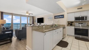 A modern kitchen with white cabinets, a granite countertop, and stainless steel appliances. There is a dining area with a gray sofa and a ceiling fan. A large window offers a view of the ocean. - FREE Beach Service & Perks! Emerald Beach 732 Pool, Hot Tub, Starbucks, Gym! (Panama City Beach)