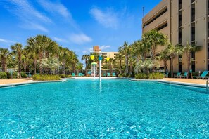 A clear blue pool surrounded by palm trees and lounge chairs, with a building in the background. - FREE Beach Service & Perks! Emerald Beach 732 Pool, Hot Tub, Starbucks, Gym! (Panama City Beach)
