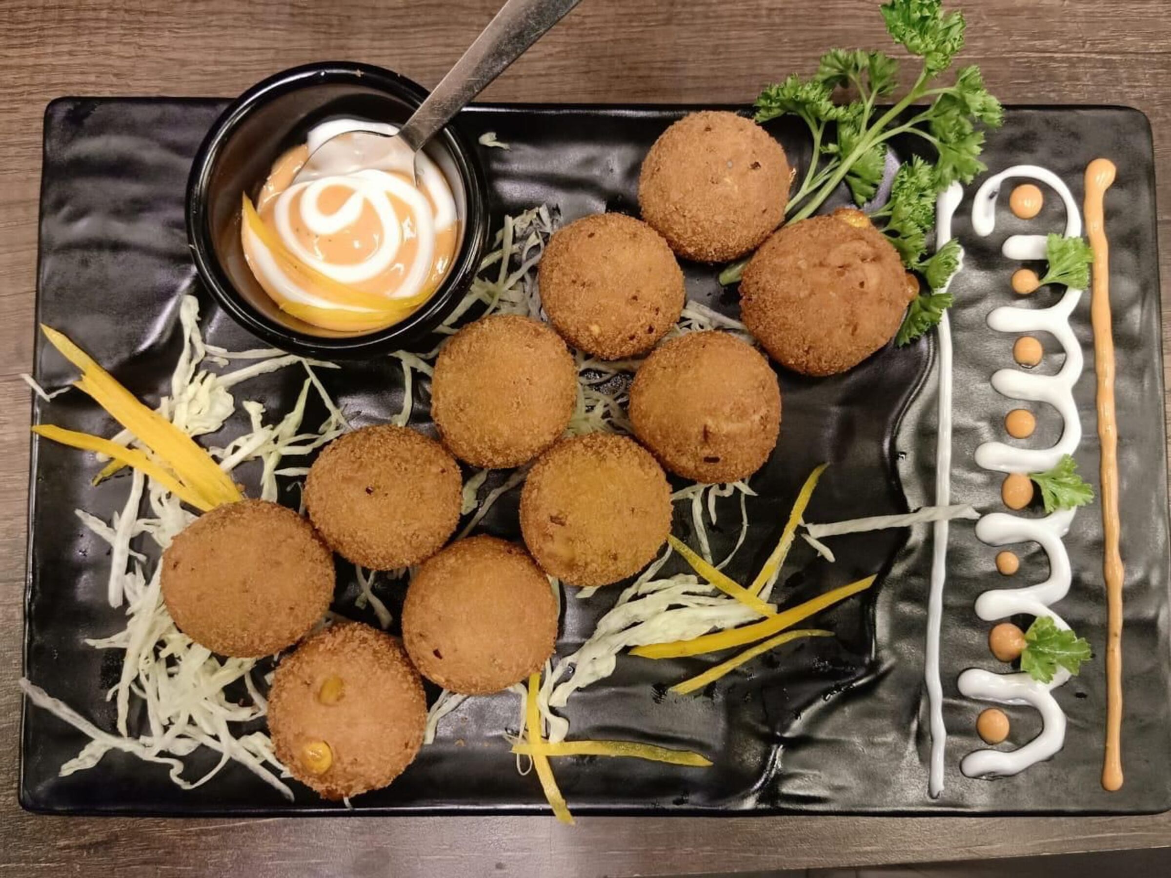 Une assiette noire contenant des croquettes, des légumes coupés en julienne et un petit bol de sauce.