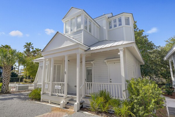 Une maison blanche de style colonial, dotée d’un porche, d’une petite structure ressemblant à une tour et d’une cour clôturée où poussent différentes plantes.