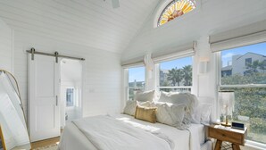 A bedroom with a large bed, a wooden nightstand, a mirror, and a view of palm trees through the window. - Lovely Beach Home, Right Across from Beach in Seaside, Fl + 2 Adult Bikes! (Santa Rosa Beach)