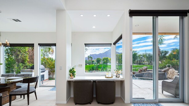 A modern kitchen with a large window, a dining area with a table and chairs, and a view of a garden and mountains. - Incredible Views From This 4 Bedroom Pool Home With Office and Sofa Sleeper (La Quinta)