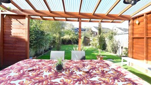Un patio cubierto con una mesa lista para comer, una planta en maceta y vistas a un jardín cuidado y casas vecinas.