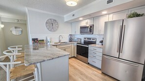 A modern kitchen with a central island, stainless steel appliances, and a large wall clock. - 2106 Brighton | On Ocean | Top Floor | Resort Access (Myrtle Beach)