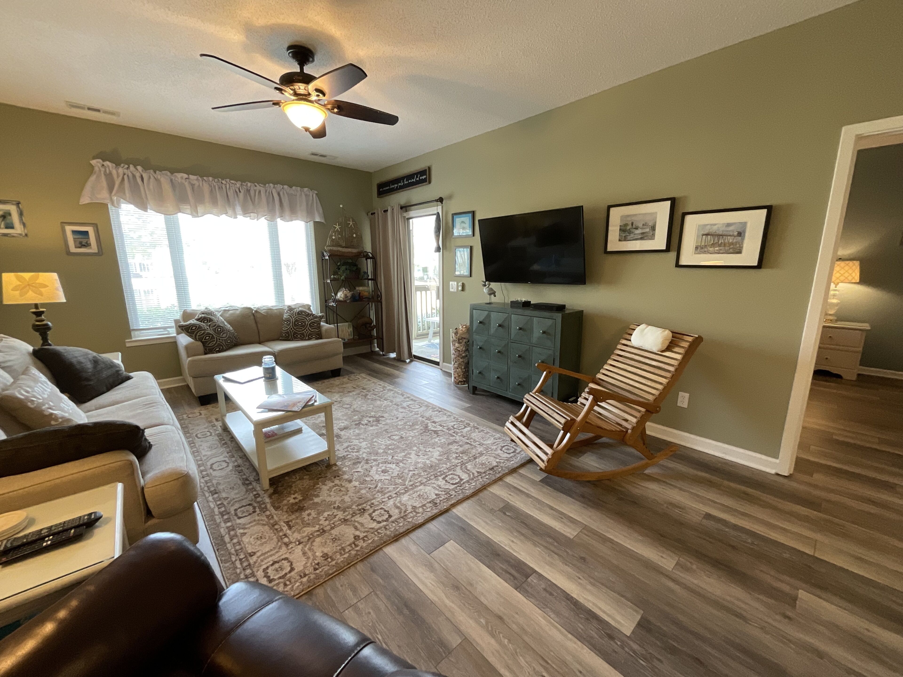 A living room with a sofa, coffee table, and a rocking chair. There is a television mounted on the wall and a ceiling fan.