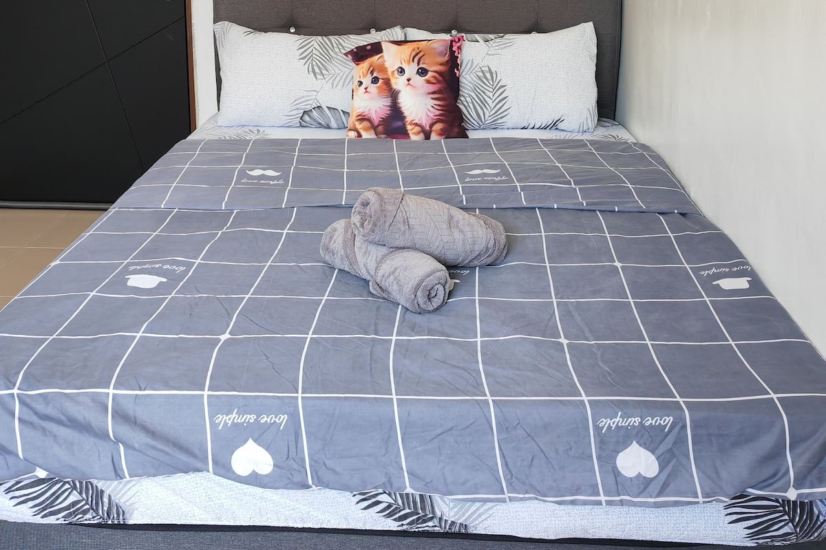 Un lit bien fait, avec une couette grise et un oreiller orné d’un motif de chat.