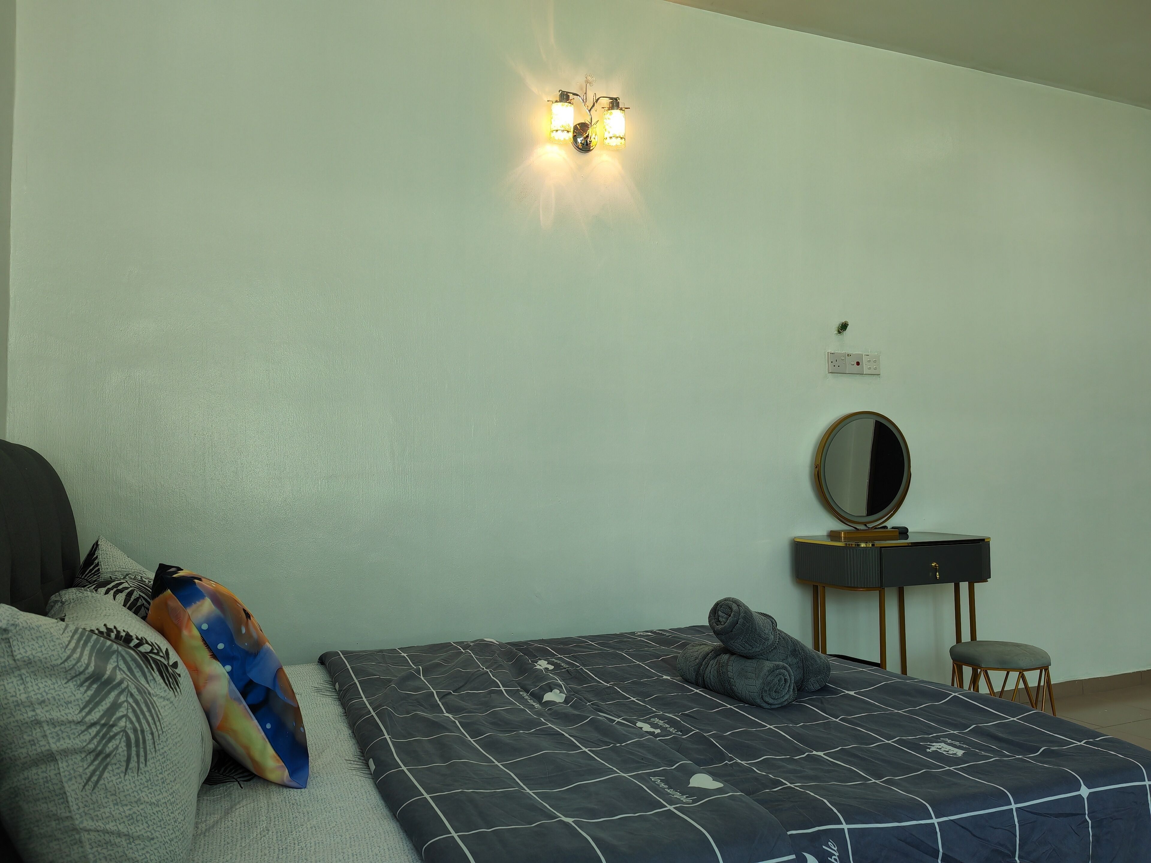 Un dormitorio con cama, almohadas, una mesita de noche, un espejo y una lámpara de pared.