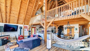 Ein großzügiger Wohnbereich im Loft-Stil mit hohen Decken, Holzbalken, einem großen Fernseher, einem Kamin, einer Hängematte und einem Essbereich.