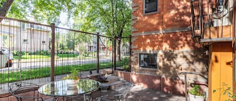 An outdoor patio with a round glass table and metal chairs, surrounded by a brick wall and a metal fence.