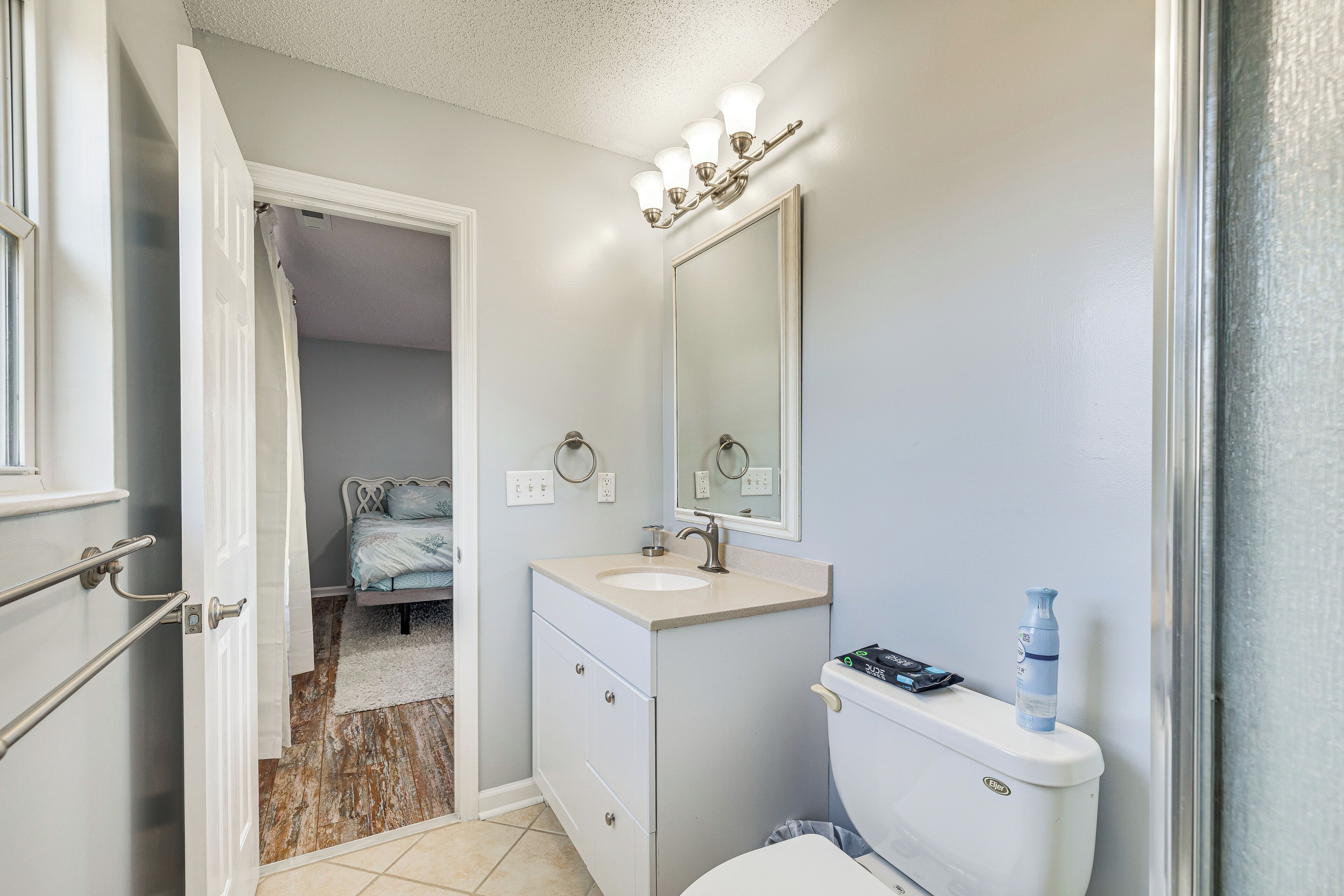 A bathroom with a toilet, sink, and mirror. There is a shower curtain partially open, revealing a bedroom with a bed and a window.