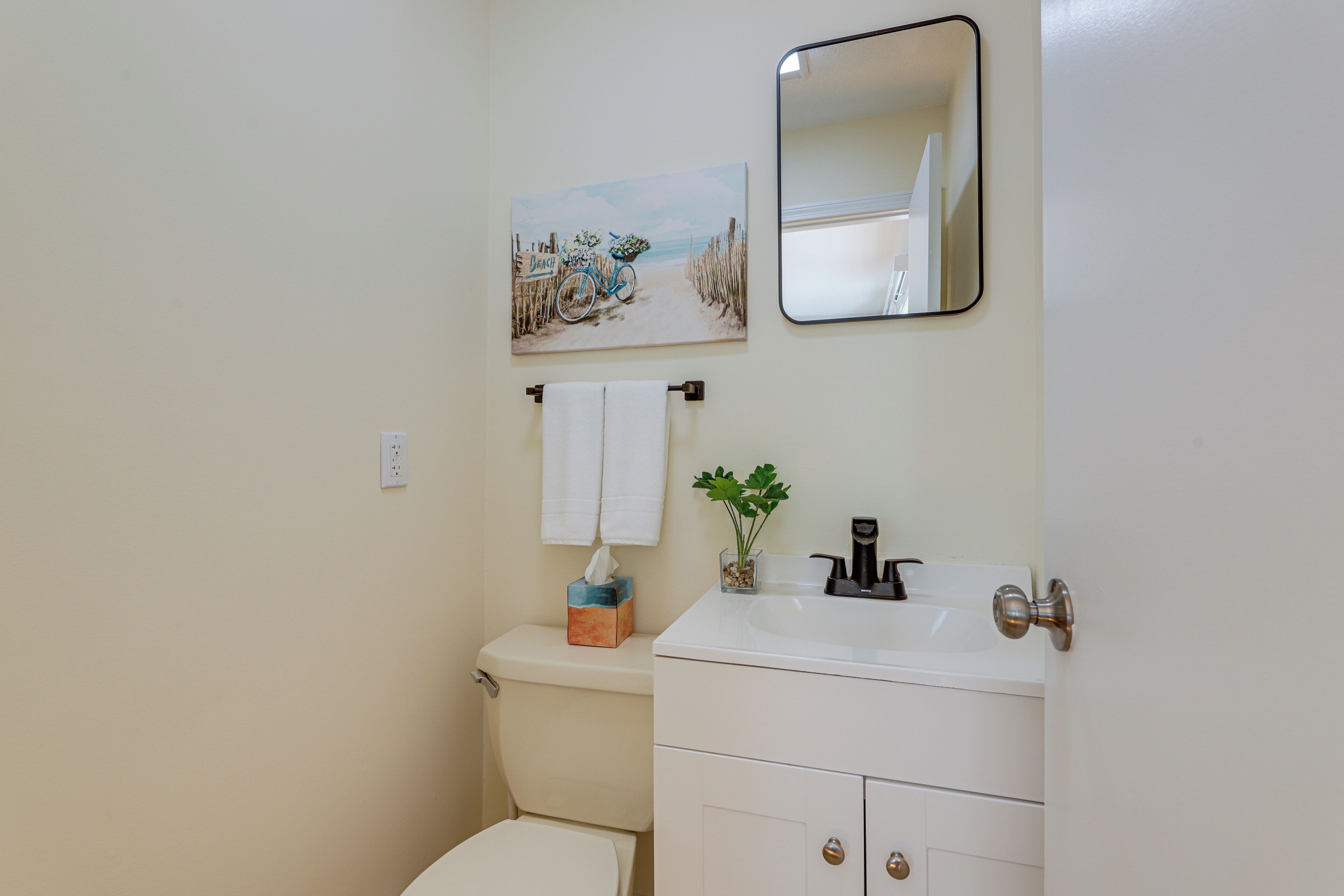 A bathroom with a toilet, sink, and a mirror. There is a towel rack with a towel and a small plant on the sink.