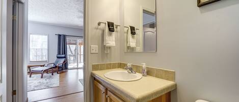 Un baño con lavamanos, espejo e inodoro. En la habitación contigua, hay una silla y una ventana.