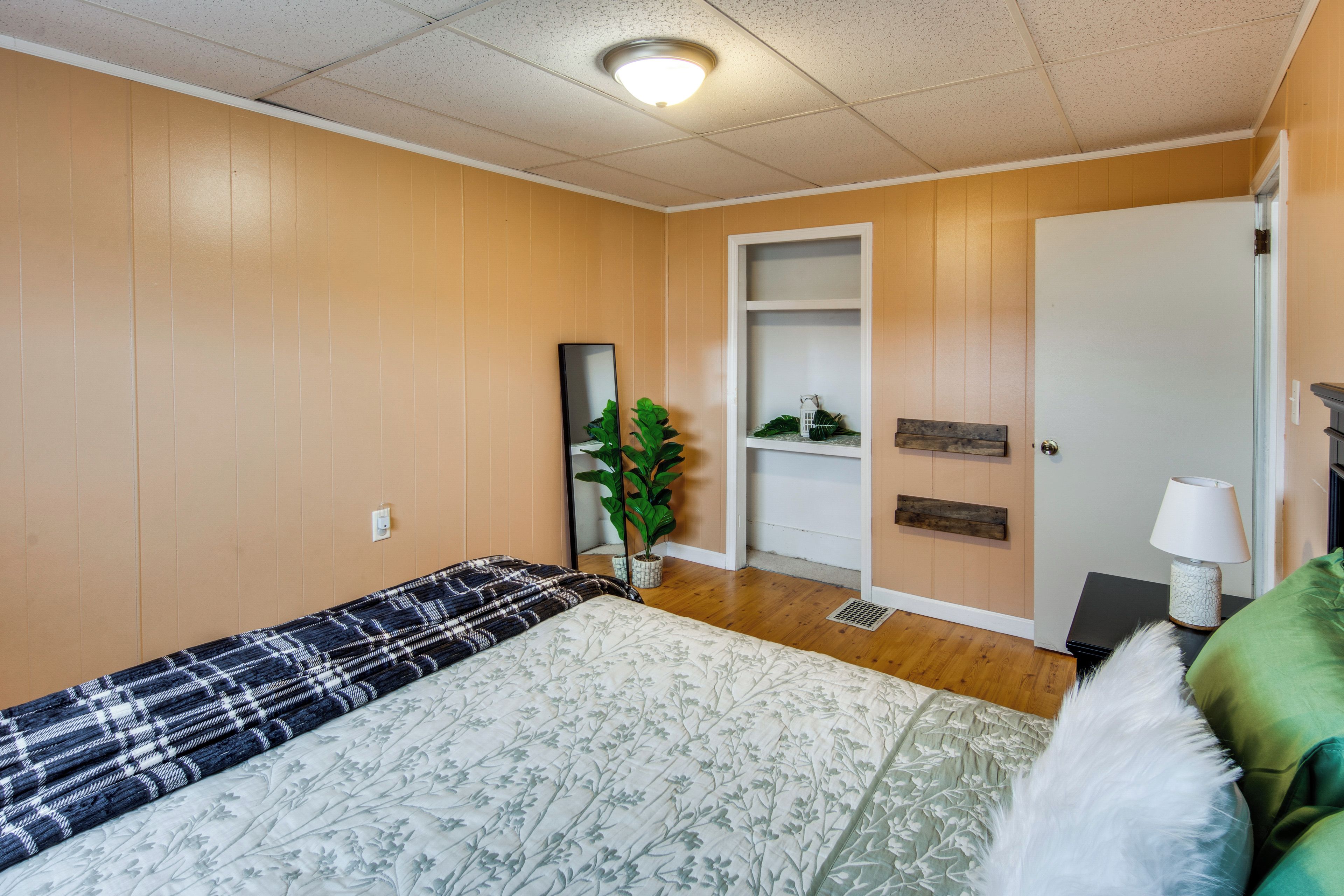 A room with a bed, a bedside table with a lamp, a potted plant, and a door leading to another room.