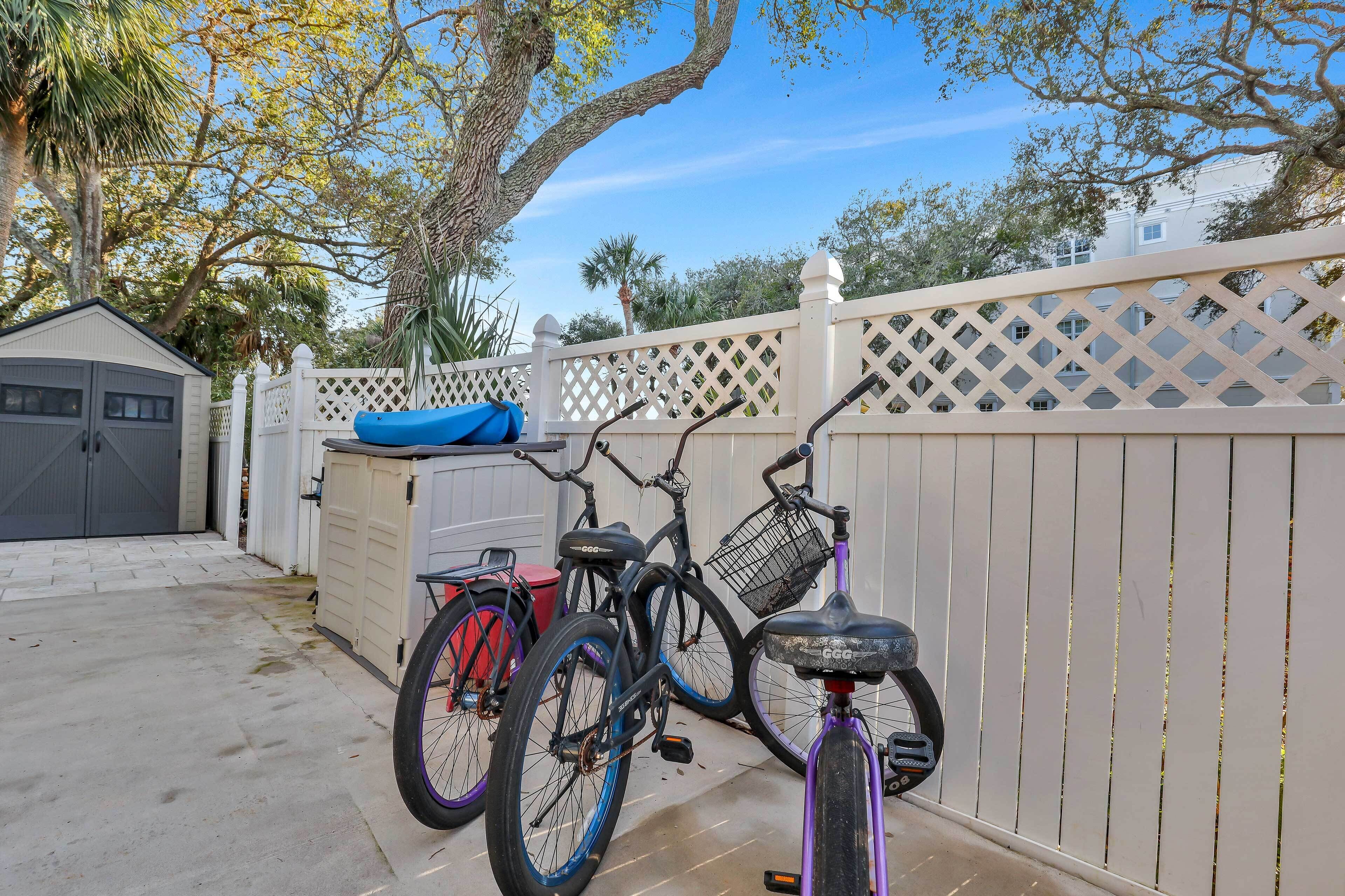 Uma bicicleta encostada em uma cerca de treliça branca, uma bicicleta vermelha, e um caiaque azul ao fundo.