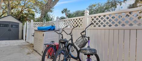 Uma bicicleta encostada em uma cerca de treliça branca, uma bicicleta vermelha, e um caiaque azul ao fundo.