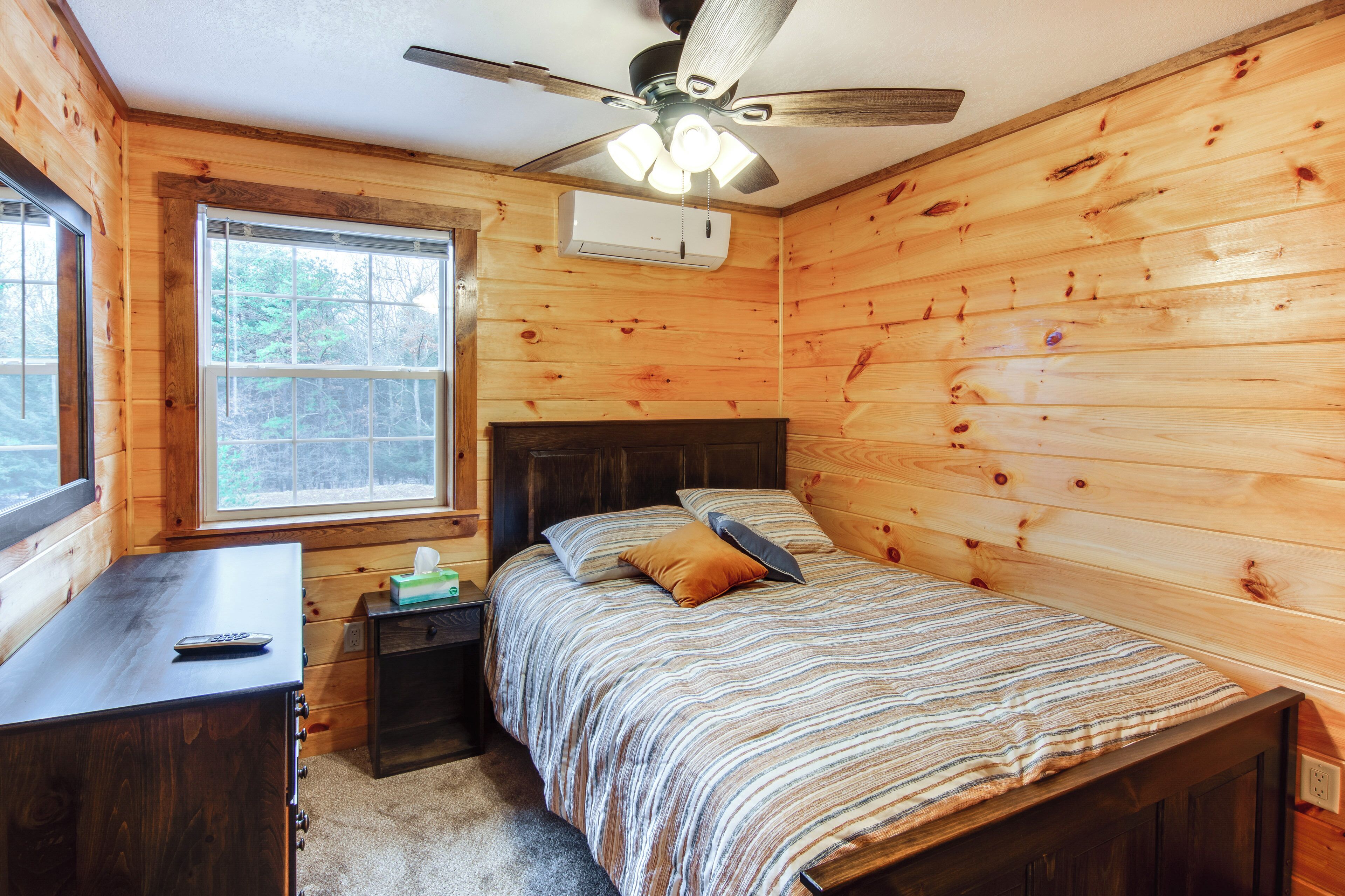 A wooden cabin bedroom with a bed, a desk, a nightstand, a ceiling fan, and a window.