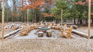 A circular fire pit surrounded by wooden Adirondack chairs in a forested area with logs and gravel ground cover. - Houtzdale Home w/ Shuffleboard & Pickleball! (Houtzdale)