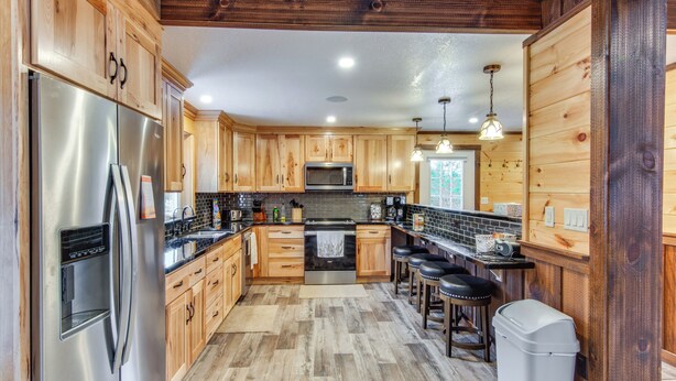 A kitchen with wooden cabinets, stainless steel appliances, and a bar area with stools. - Houtzdale Home w/ Shuffleboard & Pickleball! (Houtzdale)