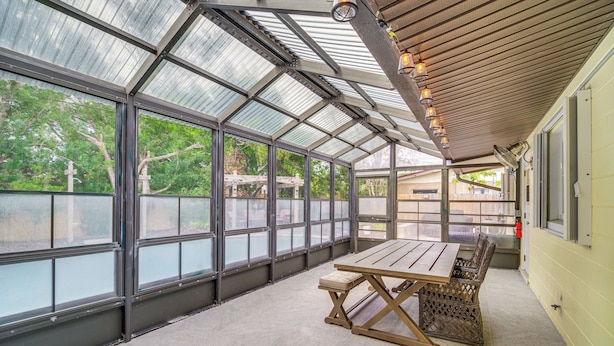 A covered patio with a table and chairs, large windows, and a view of trees.