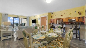 A living room with a glass dining table, chairs with pineapple patterns, a sofa, and a TV. The kitchen is visible with wooden cabinets and a microwave. - Moondrifter 207 - Condo with Spectacular Views (Panama City Beach)