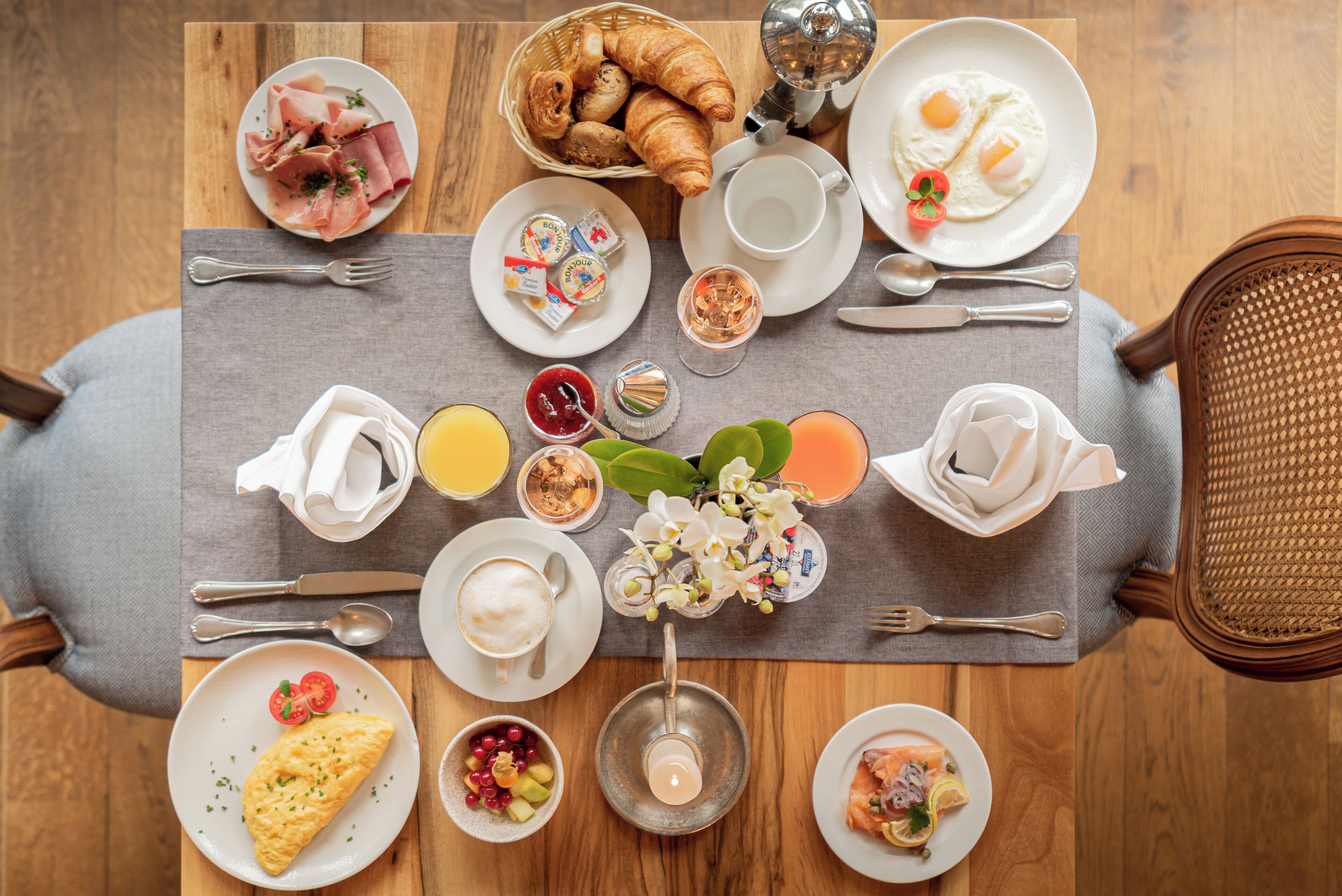 Une table en bois dressée pour le petit-déjeuner, avec des croissants, des œufs, du jambon et divers fruits.