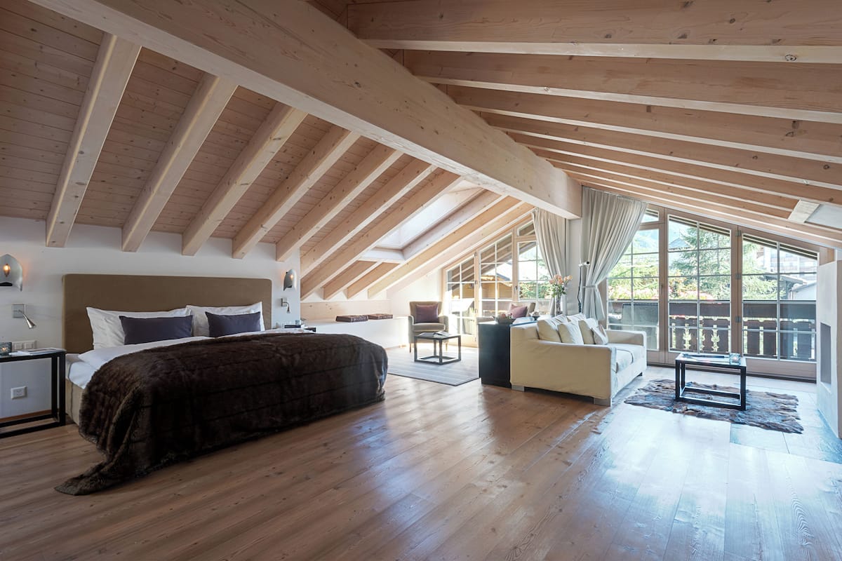 A spacious room with a large bed, a sofa, and a coffee table. The room has wooden beams on the ceiling and large windows.