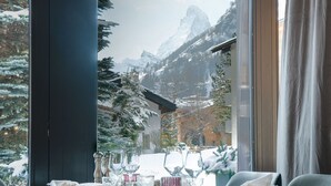 A dining area with a table set for two, a window with a view of snow-covered mountains, and a wooden ceiling. - Mirabeau Alpine (Zermatt)
