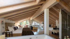 A spacious attic bedroom with a large white bathtub, wooden beams, a cozy seating area, and a glass shower. - Mirabeau Alpine (Zermatt)
