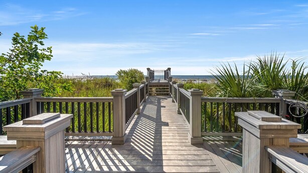 A wooden boardwalk leading to a gate, surrounded by greenery and overlooking a beach.