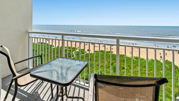 Un balcon avec une table et des chaises en verre, donnant sur une plage animée et un ciel dégagé.