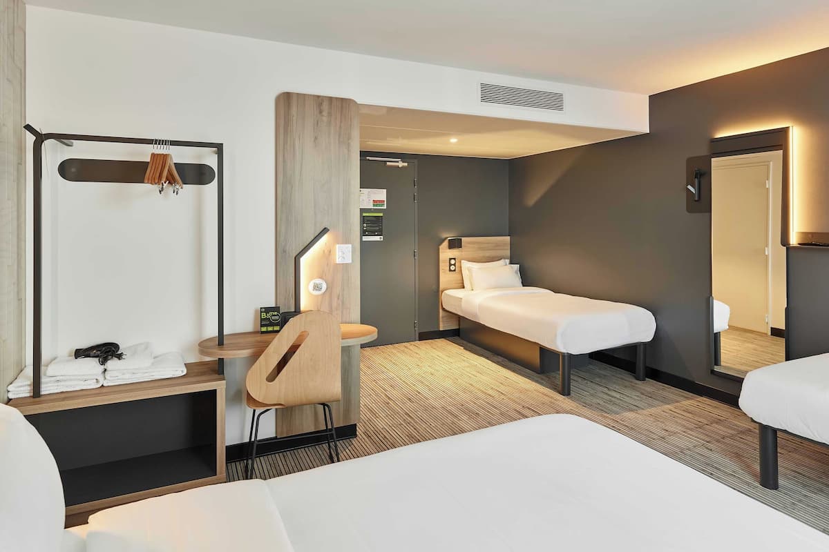Ein modernes Hotelzimmer mit zwei Einzelbetten, einem Holztisch mit Stuhl, einem Kleiderständer und einem kleinen Tisch mit Handtüchern.