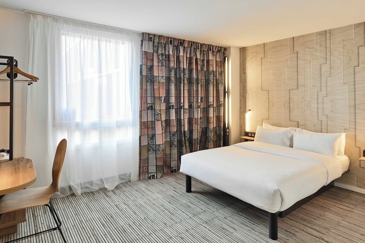 Ein Hotelzimmer mit einem großen Bett, einem Holzstuhl, einem Schreibtisch und einem gemusterten Vorhang.