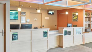 A reception desk with two computer stations, a television mounted on the wall, and a sign indicating a first aid room. - B&B Hotel Marne-La-Vallée Val D'Europe (Serris)