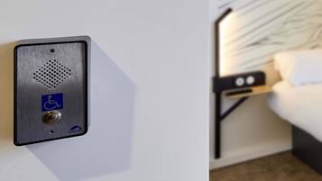 A hotel room with a bed, a nightstand, and a wall-mounted control panel with a wheelchair accessibility symbol.