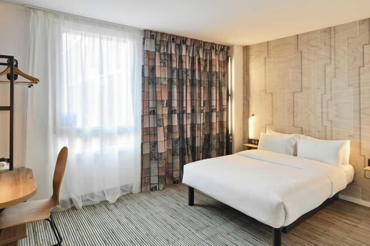 Ein Hotelzimmer mit Bett, Schreibtisch, Sessel und einem Vorhang mit Muster. An der Wand ist ein geometrisches Design zu sehen.