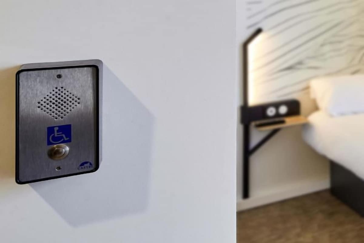 Habitación de hotel con cama, mesita de noche y panel de control en la pared con símbolo de accesibilidad para sillas de ruedas.