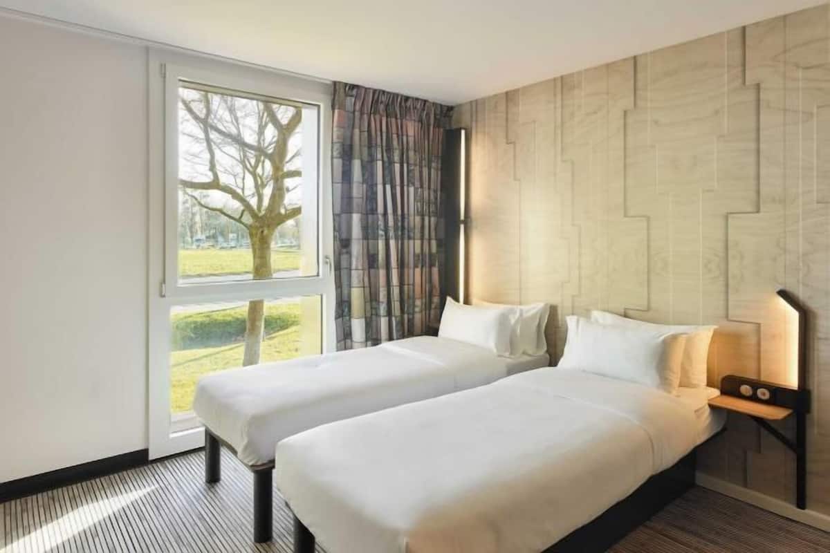 Ein Hotelzimmer mit zwei Betten, einem großen Fenster mit Blick auf einen Baum und Rasen, und einer Wand mit Muster.