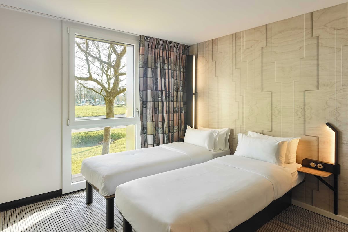 Ein Hotelzimmer mit zwei Betten, einem großen Fenster, Blick auf Baum und Gras sowie einem modernen Wanddesign.