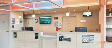A reception area with two reception desks, a clock, a digital screen, and a ceiling with geometric lighting.