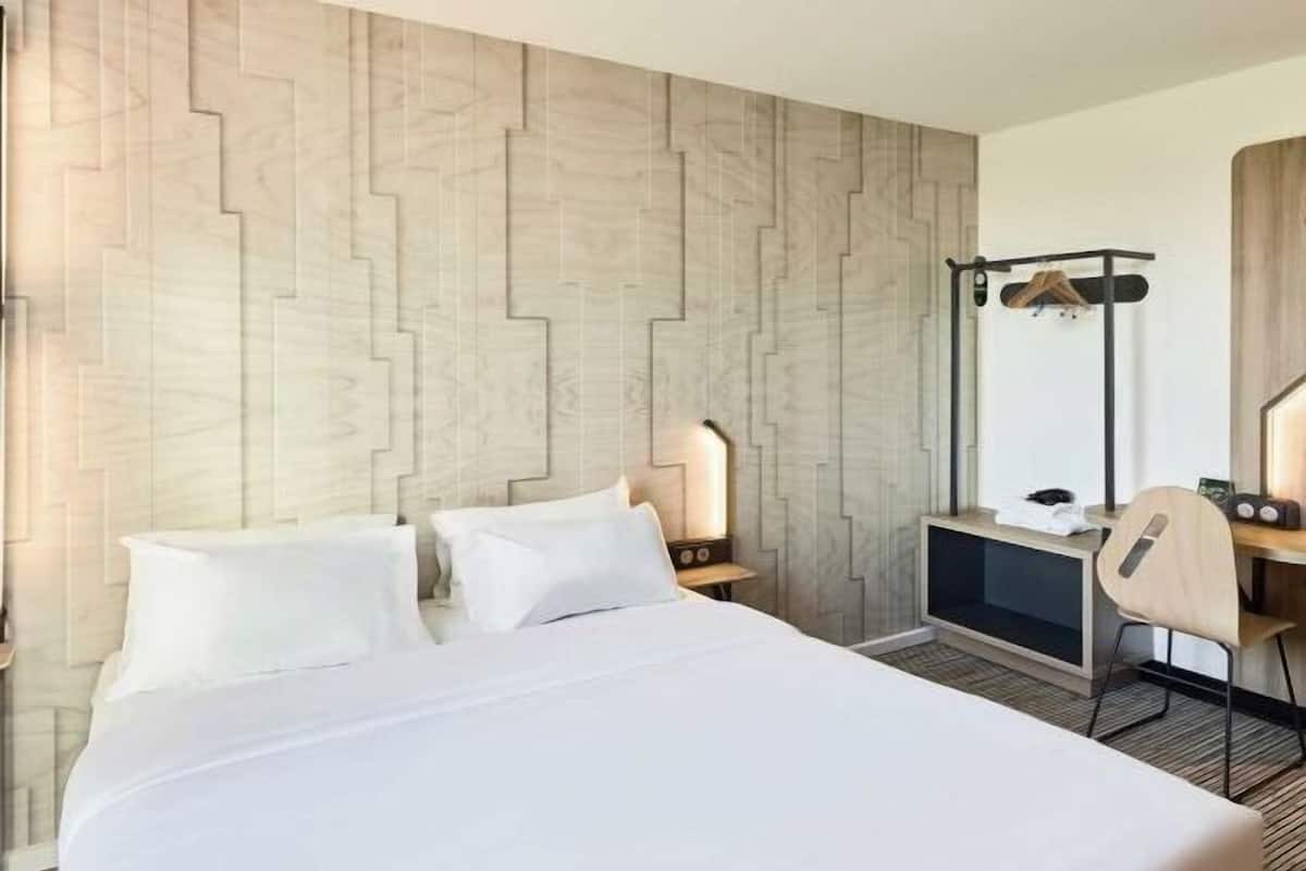 Ein Hotelzimmer mit einem Bett, einem Schreibtisch, einem Stuhl, einem Kleiderhaken und einer Wand mit geometrischem Muster.