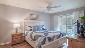Une chambre à coucher comprenant un lit, une table de chevet, une commode, un ventilateur de plafond, une fenêtre avec des rideaux et un balcon avec des chaises.