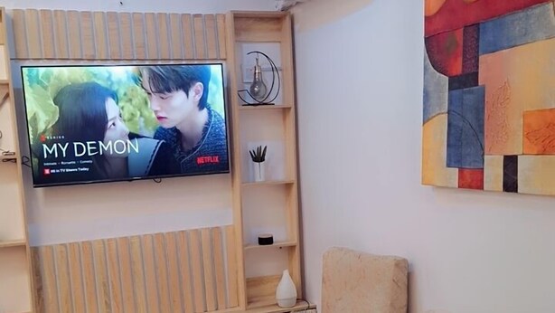 A living room with a beige sofa, a flat-screen TV mounted on a wooden shelf, an air conditioning unit, and a colorful abstract painting on the wall. - Zek Dynasty (Lagos)