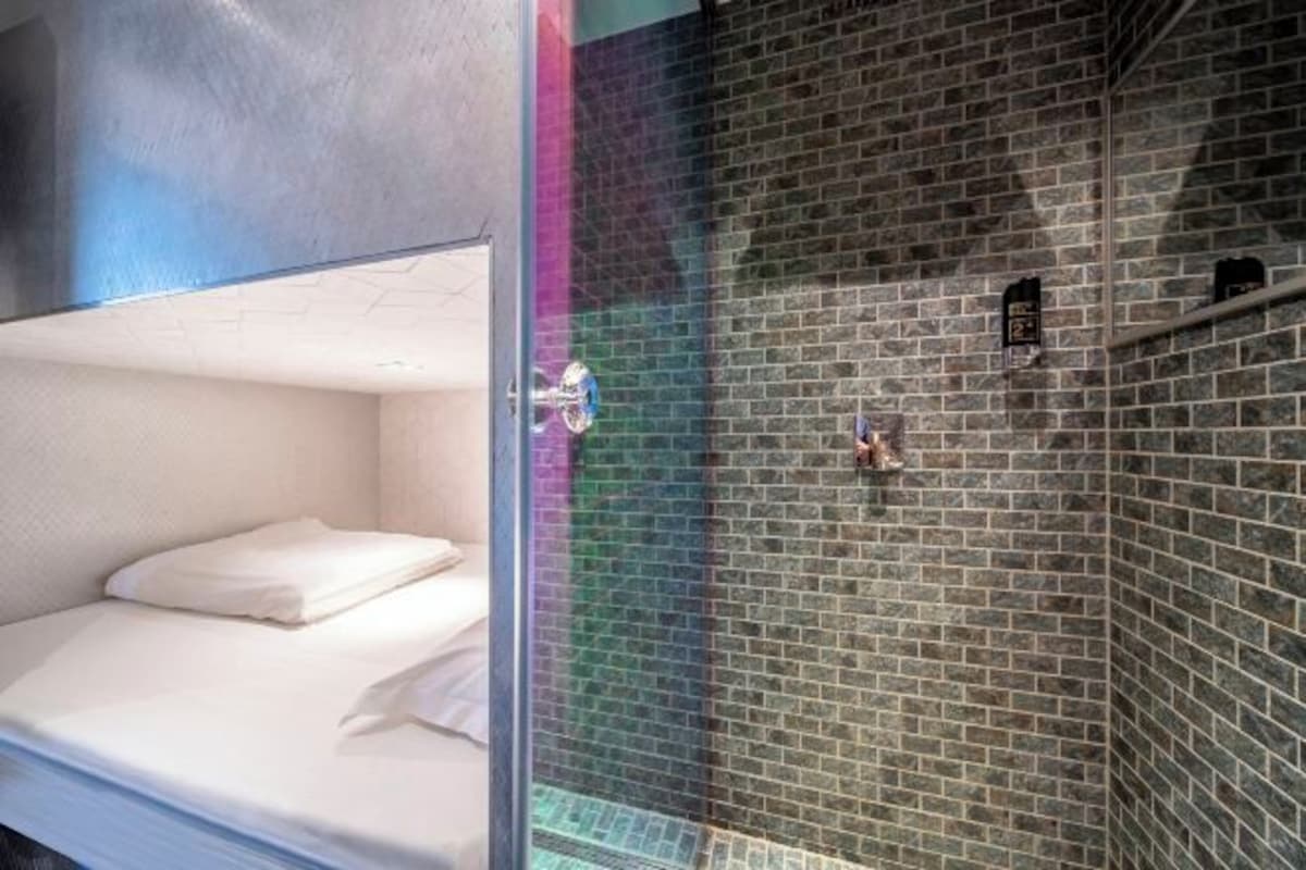 Un lit superposé avec un matelas blanc et une cloison en verre qui le sépare d’un espace douche dont le mur est recouvert de carreaux.