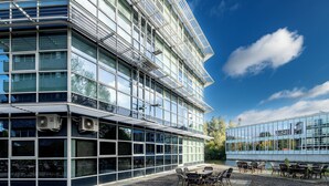 Un bâtiment moderne doté de grandes fenêtres en verre, d’un espace extérieur aménagé avec des tables et des chaises, et de climatiseurs installés à l’extérieur.