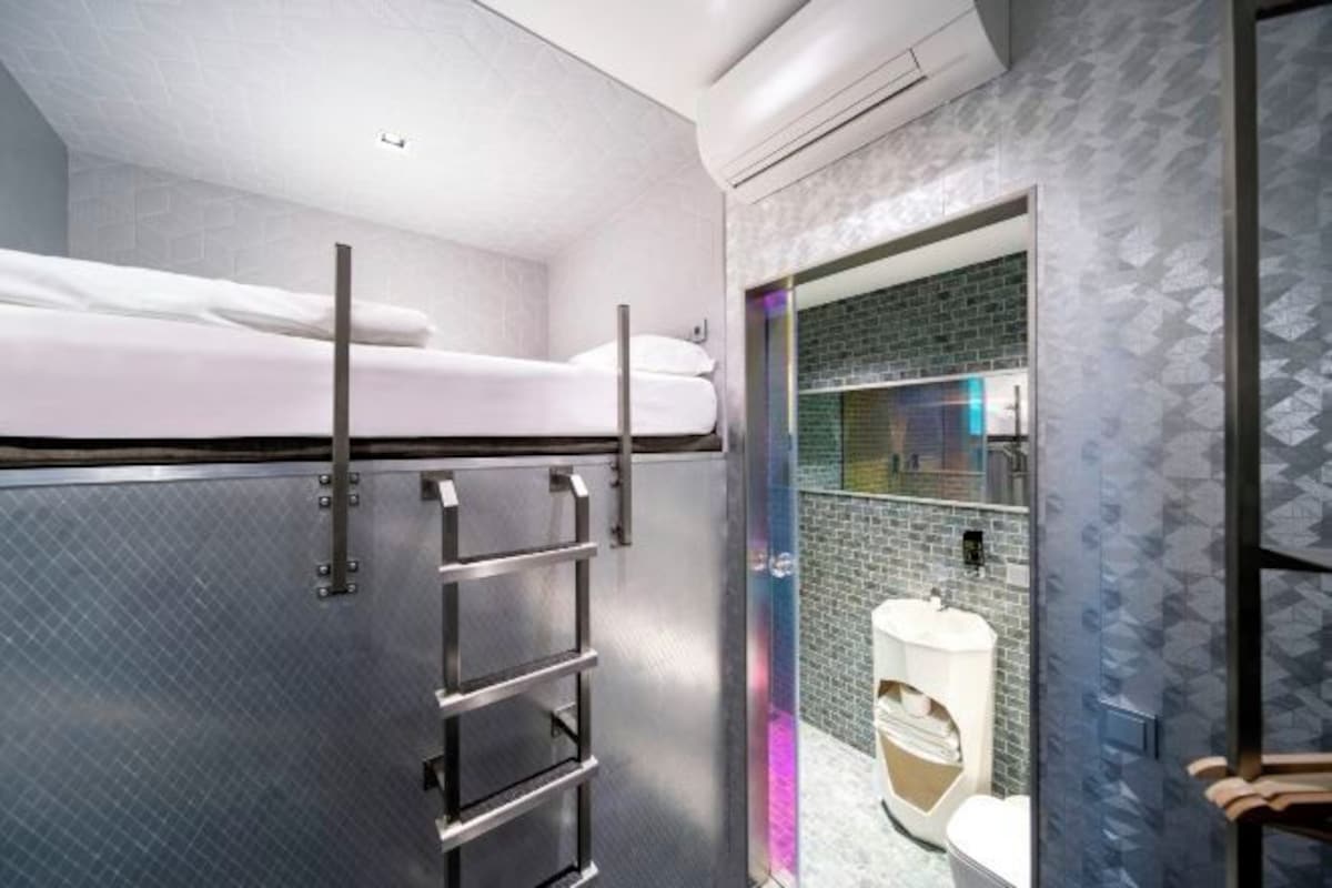 Une chambre d’hôtel compacte avec un lit superposé, une échelle en métal, une petite salle de bain avec des toilettes et un lavabo, et un mur orné d’un motif.