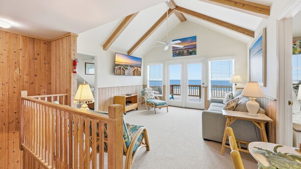 A spacious living room with a high ceiling, wooden beams, a large window with a view of the sea, a sofa, a chair, and a table with a lamp. - Arrogantly Shabby - Oceanfront (Pawleys Island)